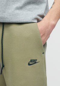 Άτομο που φοράει χακί πράσινο παντελόνι φόρμας Nike με μαύρο κορδόνι, το χέρι στην τσέπη και ένα ανοιχτό γκρι πουκάμισο χαλαρά γυρισμένο στη μέση.