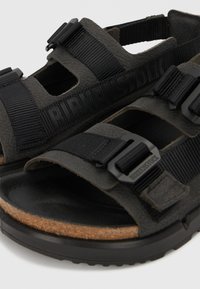 Μαύρες σανδάλια Birkenstock με ρυθμιζόμενους ιμάντες, φελλό πάτο και υφή στη σόλα, σε κοντινή λήψη με λευκό φόντο.
