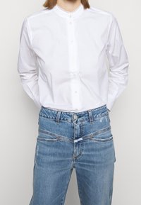 Chemise en coton blanche à manches longues avec un col montant, dotée d'une fermeture à boutons et d'une bordure en dentelle le long de l'encolure. Assortie avec un jean en denim bleu clair.