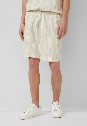 Mężczyzna ubrany w beżowo-białe paskowane szorty w zestawie z dopasowanym topem i białymi trampkami, stojący na tle jednolitego tła.