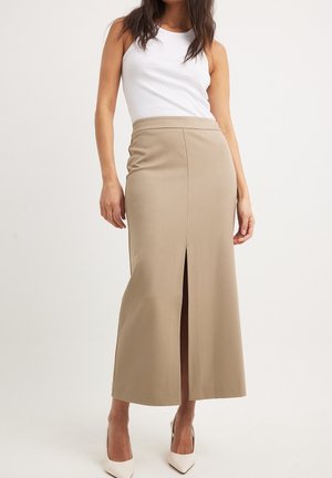 Kobieta ubrana w biały top bez rękawów, beżową spódnicę midi z rozcięciem z przodu oraz białe szpilki, stojąca na jednolitym tle.