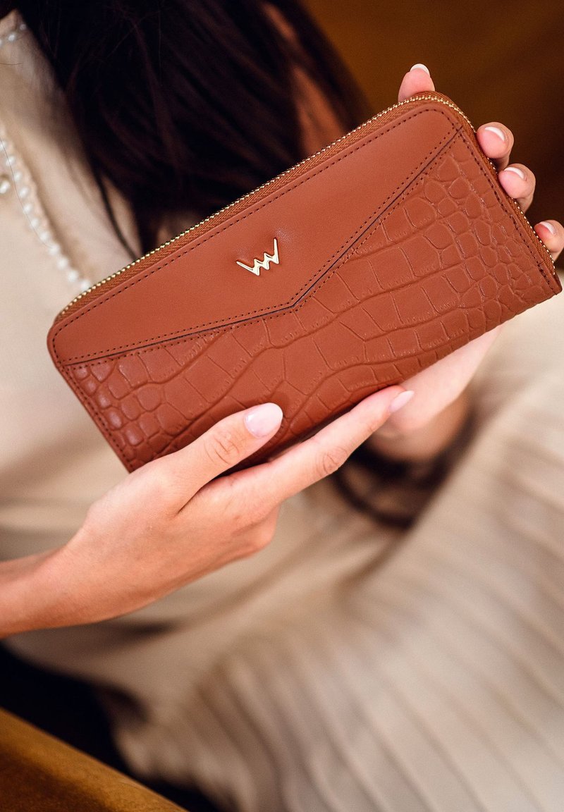 Brown crocodile-patterned leather wallet with a gold logo, zip closure, and smooth texture, held in a hand against a neutral background.
