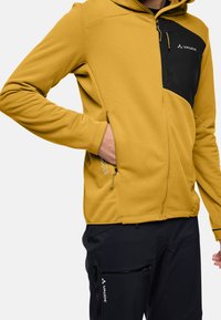 Giacca di fleece gialla con un pannello nero e cappuccio, caratterizzata da cerniere sui lati e un logo. Indossata sopra pantaloni neri con tasche laterali.