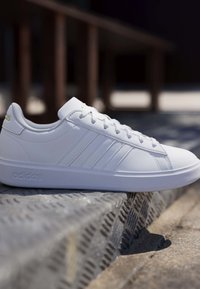 Hvid Adidas sneaker med snøredesign og tre striber på siden på en tekstureret metaloverflade i udendørs omgivelser.