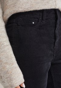 Primo piano di una mano vicino all'anca che indossa pantaloni di velluto a coste neri e un maglione di maglia beige con tasca visibile e dettagli in rivetti metallici.