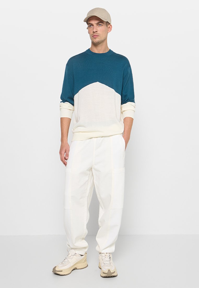 Muškarac stoji odjeven u plavu i bijelu majicu, bijele hlače, bež tenisice i bež kačket na jednostavnoj pozadini.