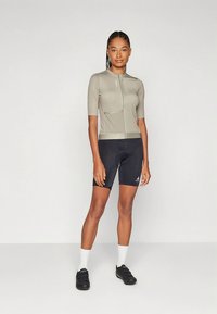 Šviesiai pilka trumpomis rankovėmis dviračių marškinėliai su priekiu užtrauktuku ir kišenėmis, derinami su juodais dviračių šortais ir sportiška juoda avalyne.