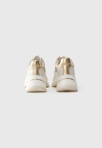 Par bež in zlatih tenisic, prikazanih s hrbtne strani na beli ozadju, ki prikazuje debele bele podplate in oznake na petah.