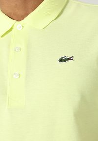 Polo tričko ve světle žluté barvě s texturovaným materiálem, tři knoflíky na předním límci a zeleným vyšívaným logem krokodýla na levé straně.