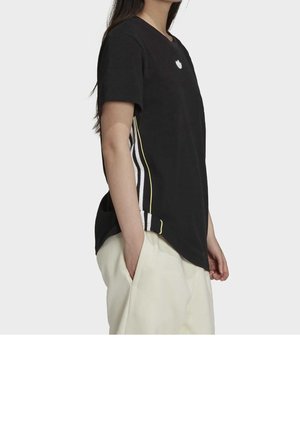 Personne portant un t-shirt noir à manches courtes avec des bandes latérales blanches et jaunes et un pantalon beige clair, les mains dans les poches, vue de côté.