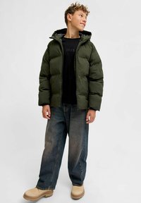 Giacca a piumino verde con cappuccio, camicia nera, jeans denim blu larghi e stivali beige. Tessuto morbido e design trapuntato visibile.