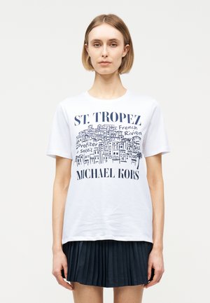 Frau trägt ein weißes St. Tropez Michael Kors T-Shirt mit maritimem Stadtbilddruck und einem schwarzen Faltenrock, stehend vor einem schlichten Hintergrund.