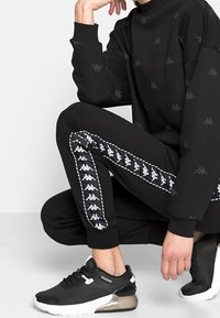 Zwarte sweatshirt en broek met subtiel logo patroon. Sweatpants hebben witte zijstrepen. Zwarte sportschoenen met gaas- en transparante accenten.