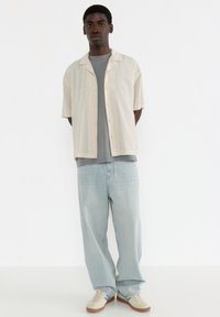 Beige Kurzarmhemd über einem grauen T-Shirt, kombiniert mit hellblauen voluminösen Jeans und beige Sneakern mit blauen Akzenten.