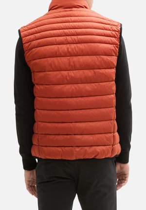 Orange puffervest med vertikalt quiltet design og høj krave, båret over en sort langærmet skjorte, med en glat, skinnende finish.