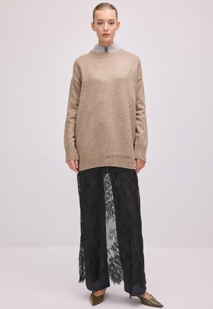 Femme portant un pull beige oversize par-dessus une chemise grise colossale et une jupe en dentelle noire transparente avec des talons pointus, debout devant un fond uni.