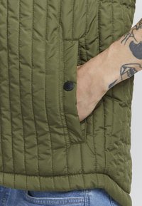 Chaleco acolchado verde oliva con costuras horizontales, un bolsillo lateral con cierre a presión negro y un dobladillo recto.