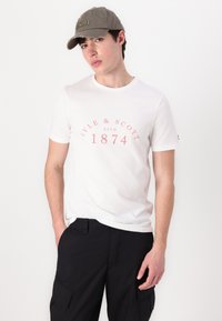 Tricou alb din bumbac, cu mâneci scurte și detalii cu un logo roz; purtat cu pantaloni cargo negri și o șapcă gri cu text brodat.