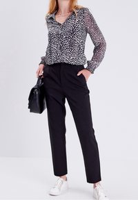 Durchsichtige Bluse mit schwarz-weißem Leopardenmuster, langen Ärmeln mit elastischen Bündchen, kombiniert mit schwarzen eleganten Hosen und weißen Sneakers.