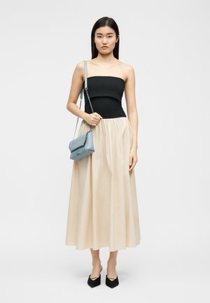 Moteris stovi, vilkinti juodą be petnešėlių viršutinę dalį ir ilgą smėlio spalvos sijoną, laiko šviesiai mėlyną peties krepšį, dėvi juodus smailius batus, neutralus fonas.