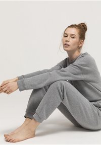 Grijze katoenen sweatshirt en joggers set, met ribgebreide manchetten, een ontspannen pasvorm en een zachte textuur. Model zit met blote voeten.