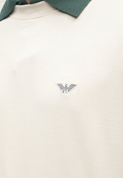 Gaļējs gaiši bēšs polo krekls ar teksturētu zaļu apkakli, pie krūšu zonas neliels izšūts balts un tumši zils logo.