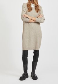 Robe-pull beige en tricot avec poignets et ourlet côtelés, associée à un pantalon noir ajusté et des bottes noires montant jusqu'au genou. Texture lisse, coupe décontractée.