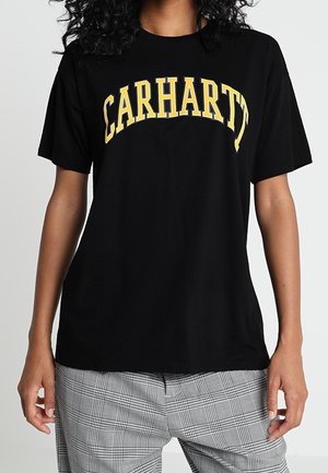 Black cotton t-shirt with yellow "CARHARTT" text printed in an arched design. Short sleeves, relaxed fit, paired with plaid trousers.