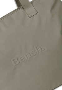 Sac fourre-tout en tissu vert avec un logo "Bench." en relief à l'avant. Livré avec une poignée assortie et résistante. Texture lisse, design minimaliste.