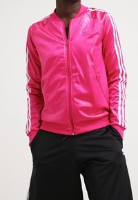 Klart rosa satin bomberjacka med vita ränder på ärmarna, dragkedja och ribbat halsband, kombinerad med svarta träningsbyxor.