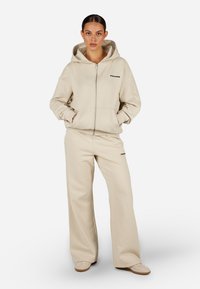 Bézs cipzáras kapucnis pulóver nagy kapucnival és elülső zsebekkel, hozzá illő, bő szárú nadrággal párosítva. Finom logó részletekkel díszítve.