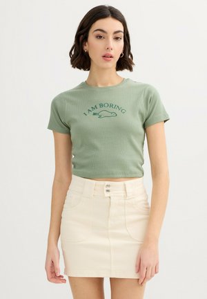 Mujer con cabello corto y oscuro que lleva un top corto verde con el texto "I AM BORING" y una minifalda color crema con botones, de pie contra un fondo blanco.