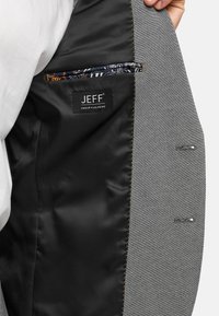 Unutrašnjost sivoj jakne s crnim podstavom. Sadrži oznaku JEFF Urban Tailoring, dva uzorčena džepa i detalje od žutog šivanja.