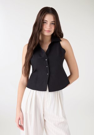 Giovane donna con lunghi capelli castani, che indossa un top nero senza maniche con bottoni e pantaloni bianchi a righe con gambe larghe, in piedi su sfondo bianco.