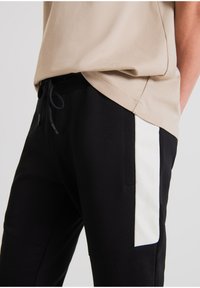 Zwarte joggers met een contrasterend wit zijpaneel, elastische tailleband met trekkoord, en zachte, gestructureerde stof. Beige top heeft een relaxte pasvorm.