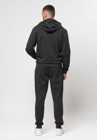 Sötét szürke kapucnis felső, kiemelkedő hátsó kapucnival, hozzáillő melegítőnadrággal párosítva. Puha, textúrált anyaggal és laza szabással rendelkezik. Fehér sportcipők.