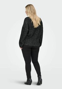 Blonda sieviete stāv ar muguru vērsta, tērpusies melnā raibā džemperī, melnās šaurās biksēs un melnās apavos ar papēžiem.