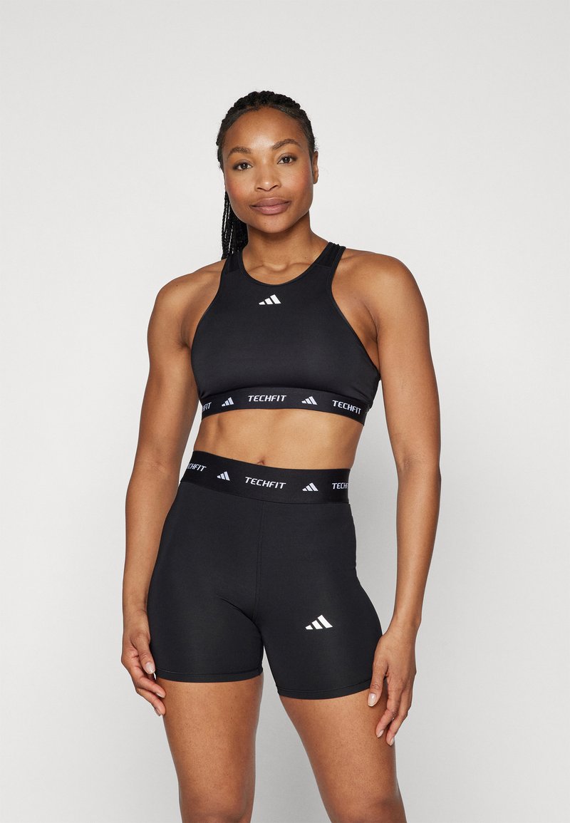 Svart Adidas Techfit sports-BH og shorts, med subtile hvite logo-detaljer og en tettsittende passform. Materialet ser glatt og elastisk ut.