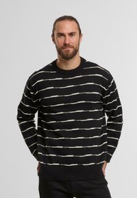 Maglione nero con righe orizzontali bianche, texture lavorata a maglia, colletto rotondo e polsini e orlo a coste, indossato da un modello su uno sfondo semplice.