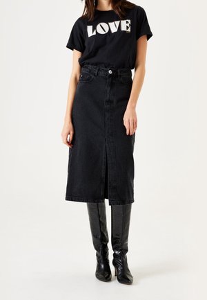 Vrouw draagt een zwarte "LOVE" t-shirt, zwarte high-waist midi spijkerrok met een split aan de voorkant, en zwarte leren laarzen tot aan de knie.