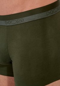 Bañador boxers en color verde oliva, fabricado en tejido suave. Presenta una cinturilla gris con el logo "HOM" en relieve. Diseño sencillo y forma ajustada.