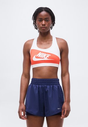 Donna atletica con dreadlocks corti che indossa un reggiseno sportivo Nike bianco e arancione e pantaloncini da corsa Nike blu navy, in piedi davanti a uno sfondo bianco.