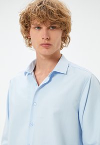 Camicia blu chiaro con bottoni, dal tessuto morbido, maniche lunghe e colletto classico. Presenta bottoni blu e una vestibilità comoda.