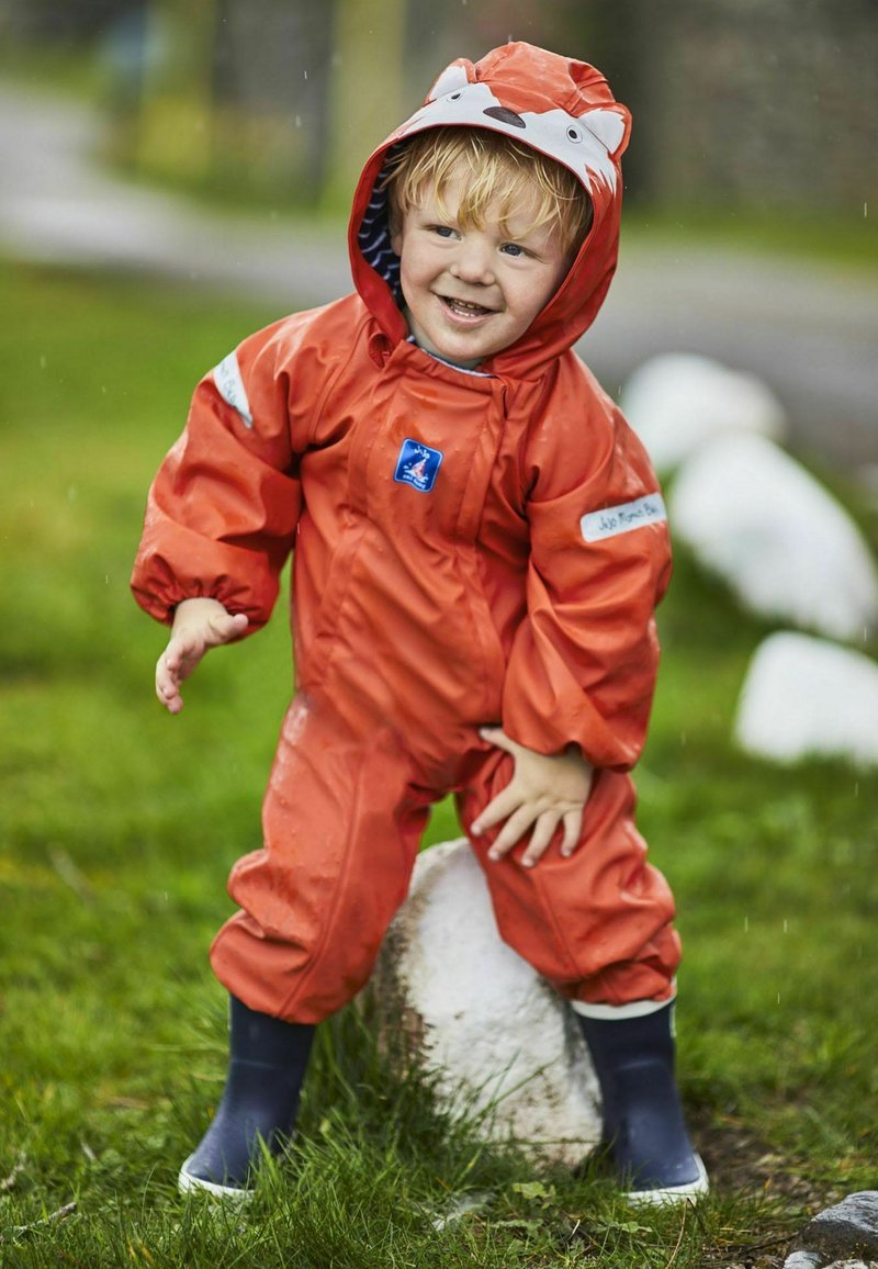 Un ensemble de pluie imperméable orange vif avec une capuche en forme de tête de renard, des poignets élastiques et des bottes en caoutchouc bleues, se tenant sur une pierre dans une zone herbeuse.
