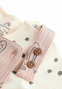 Rosa ribbade remmar med bruna logotyper fäster vid en ljus krämfärgad bodysuit med grå prickar och gröna abstrakta mönster.