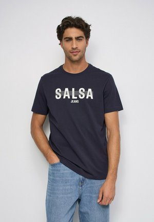 Hombre con cabello rizado oscuro que lleva una camiseta azul marino con el texto "SALSA JEANS" y vaqueros azul claro, de pie frente a una pared lisa y clara.