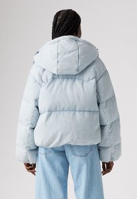 Light blue puffer jacket with a hood, oversized shape, quilted texture, and elastic cuffs, worn over matching light denim jeans.