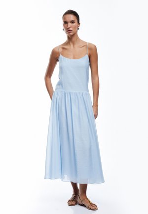 Vestido maxi sin mangas de color azul claro, con un corte holgado, que presenta una cintura fruncida y una textura suave. Llevado con sandalias planas color canela.