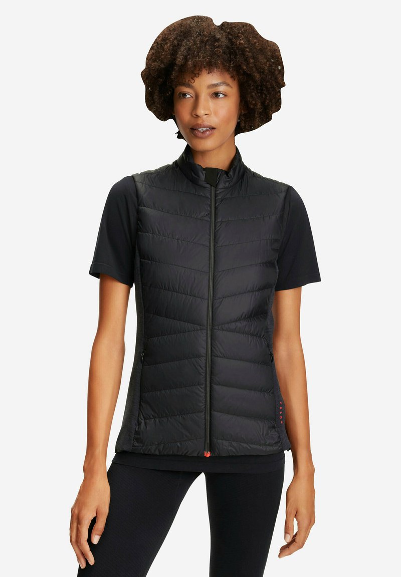 Donna con capelli ricci che indossa un gilet nero trapuntato senza maniche sopra una maglietta nera a maniche corte, in piedi davanti a uno sfondo bianco.