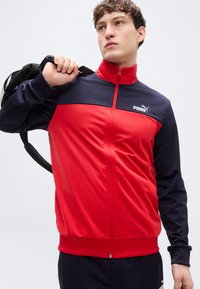 Αθλητικό τζάκετ με φερμουάρ σε κόκκινο και μπλε ναυτικό χρώμα με ψηλό γιακά, λεία υφή, λογότυπο PUMA στο αριστερό στήθος και ριμπ μανσέτες. Μαύρο σακίδιο στην πλάτη.